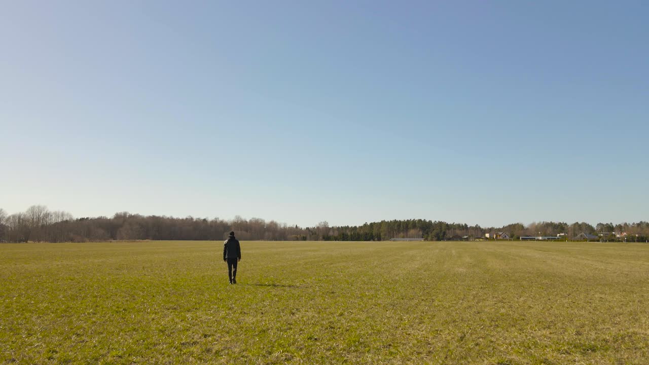 Aerial drone footage following and flying over a man who is walking further in a green and brown sunny farm field of grass field landscape during a sunny day with horizon and forest in the background
