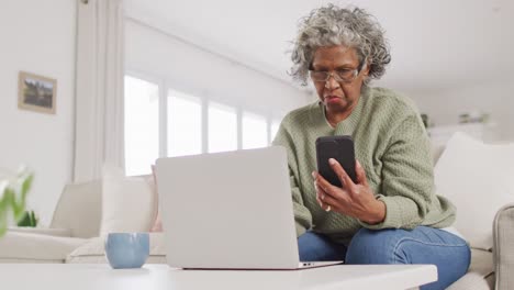Portrait of senior african american woman sitting on sofa, using laptop and smartphone