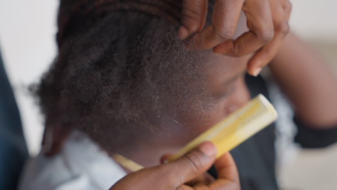 Closeup combing black childs scalp gently. Caregiver inspects hair, parts afro, uses finetooth comb to lift visible flakes and dust, soothes child between passes, warm household lighting, focused