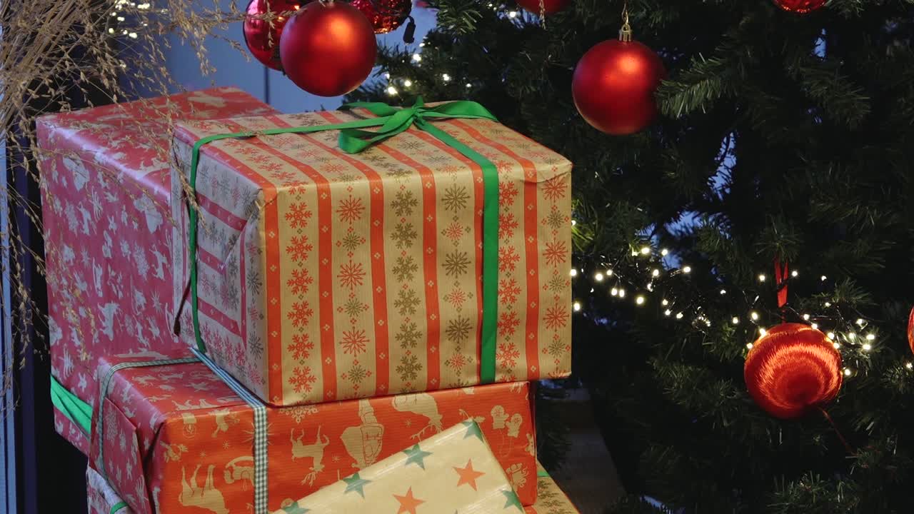 Noel temalı kağıtlar ve kurdelelerle sarılmış bir hediye yığını, kırmızı toplarla ve parıldayan ışıklarla süslenmiş aydınlatılmış bir ağacın yanına yerleştirilmiş.