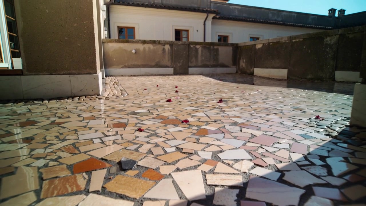 Telecamera che si allontana mostrando un pavimento a mosaico da un'angolazione bassa, con petali rossi, un parapetto in cemento e finestre.