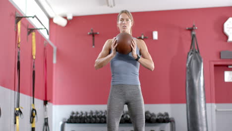 Fit-Junge-Kaukasische-Frau-Hockt-Mit-Einem-Medizinball-Im-Fitnessstudio