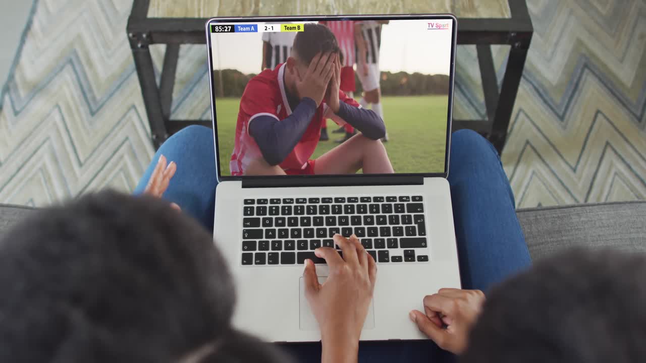 video de dos personas afroamericanas sentadas en el sofá y viendo un partido de fútbol en una computadora portátil