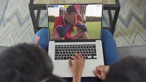 Video-Von-Zwei-Afroamerikanern,-Die-Auf-Der-Couch-Sitzen-Und-Sich-Ein-Fußballspiel-Auf-Dem-Laptop-Ansehen