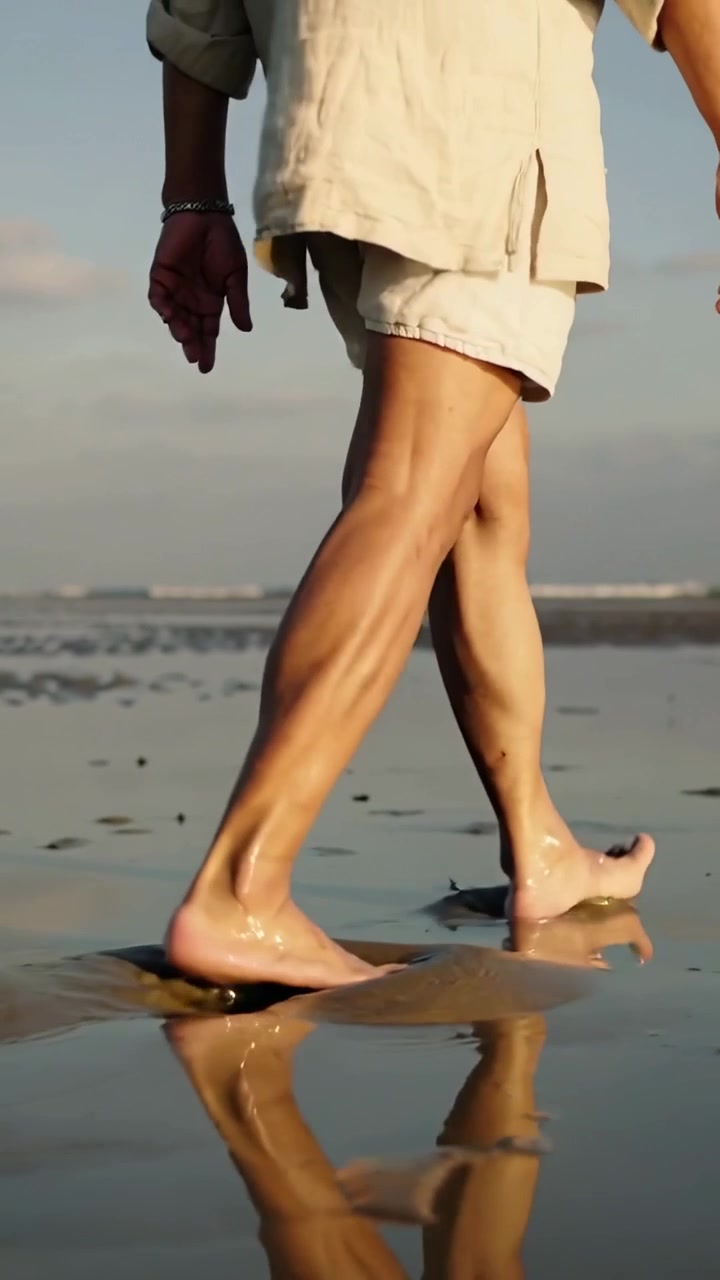 Video verticale: Uomo che cammina su una piana di marea riflettente, indossando pantaloncini chiari e un braccialetto