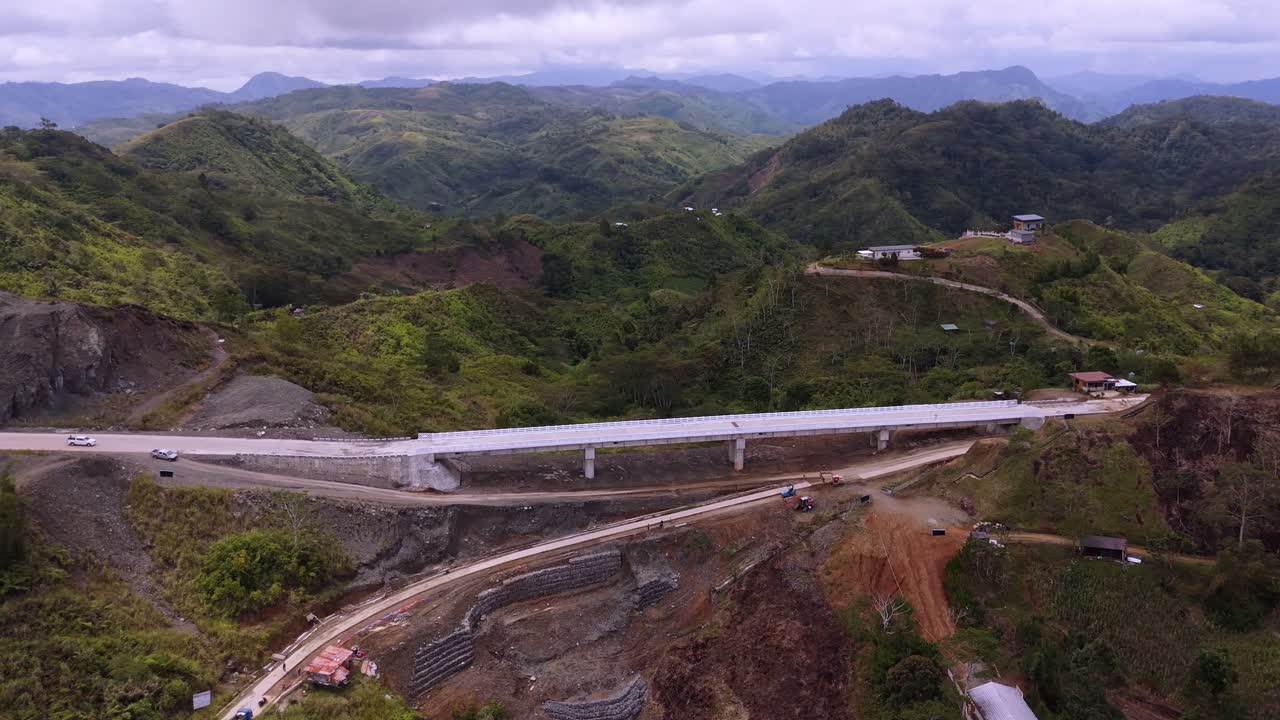 4K aerial drone side profile of a concrete bridge viaduct under construction. Shows support pillars, retaining walls (gabions) for slope protection, and unpaved service roads in a rural mountain area