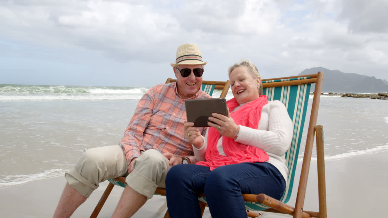 Front view of old caucasian senior couple using digital tablet and sitting on sunlounger a beach 4k