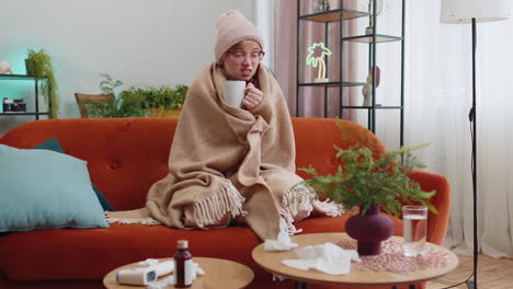 Sick-young-woman-wear-hat-wrapped-in-plaid-sit-alone-shivering-from-cold-on-sofa-drinking-hot-tea