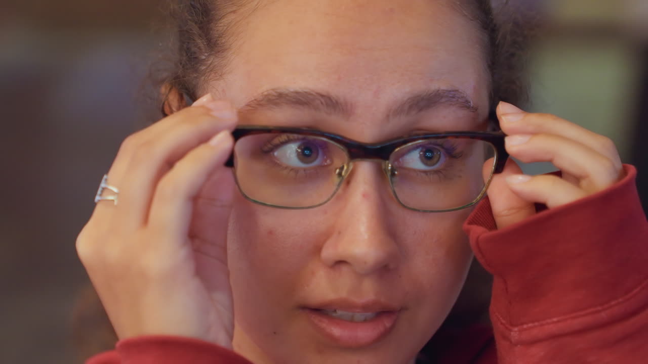 Mulher latina testando óculos com reflexos azuis, sorriso confiante e ajuste focado, armações em close-up capturam a redução de brilho da tela e clareza óptica, atmosfera que mescla showroom e café.