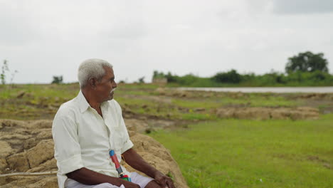 an elderly indian man sits on a rock in a rural field