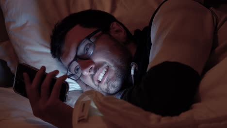 hombre acostado en la cama con un teléfono inteligente, la pantalla ilumina la cara en un dormitorio oscuro, desplazándose, redes sociales, enviando mensajes de texto a amigos en la noche antes de dormir