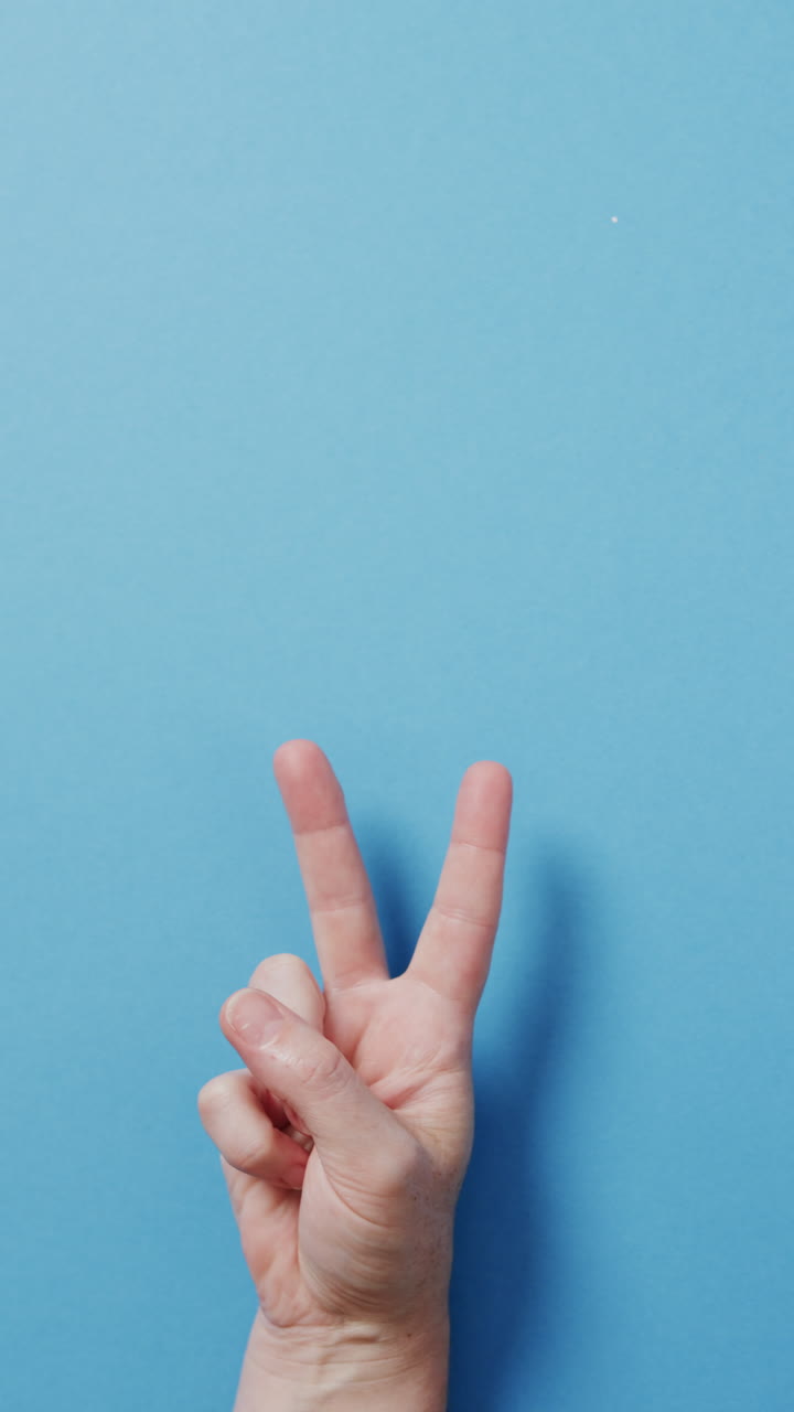 Vertical video of hand of caucasian man showing peace sign with copy space on blue background
