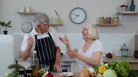 happy senior couple dancing while cooking together in kitchen with fresh vegetables and fruits