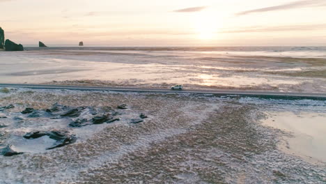 Aerial-view-orbiting-vehicle-on-Reynisfjara-main-road-travelling-across-snowy-sunlit-jagged-black-sand-beach