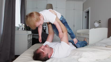 papá mantiene una dignidad por encima de sí mismo acostado en la cama. un niño con una camiseta blanca se ríe y sonríe de jugar con su padre