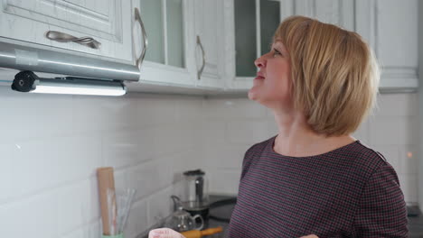 cheerful elegant woman dancing in front of kitchen cabinet, wearing plaid clothing, illuminated by under-cabinet lighting, enjoying moment in modern white kitchen with organized space