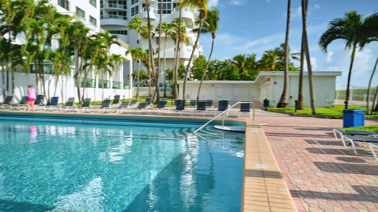Swimming pool side with lounge chairs and palm trees. Clean blue water of a resort pool with sunbeds on a brick patio and white buildings in the background during daytime