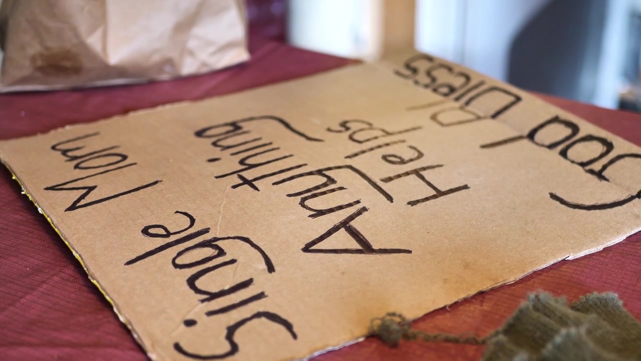 Homeless women tracing over and making cardboard sign saying, "Single mom, anything helps, God bless".