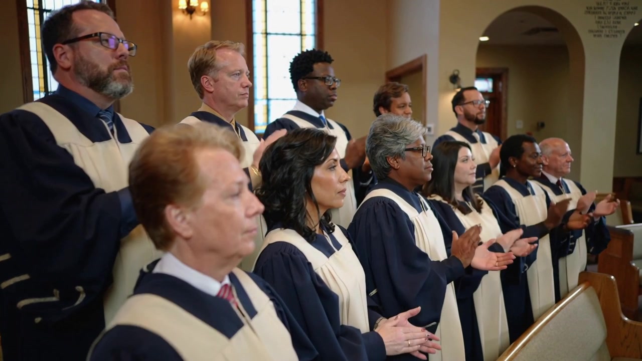Clapping choir following musical cue in church sanctuary, wearing navy robes with white stoles