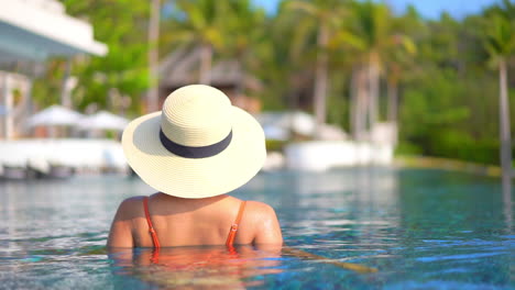 Vista-Trasera-De-Una-Modelo-Bastante-Joven-Irreconocible-En-Traje-De-Baño-Y-Sombrero-Para-El-Sol-De-Pie-Dentro-De-La-Piscina-Del-Resort-De-Isla-Tropical-En-Bali,-Indonesia,-Palmeras-Borrosas-En-El-Fondo