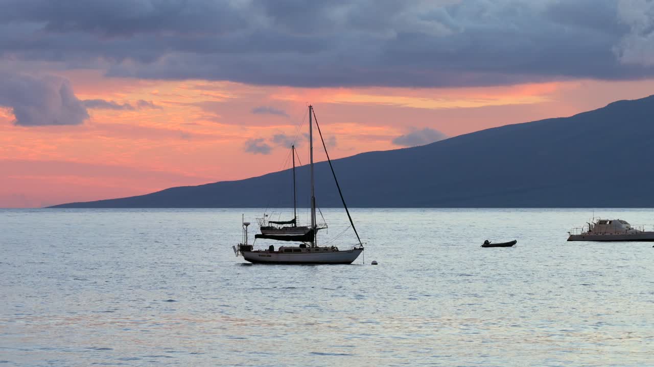 Visuale ravvicinata di pescherecci al largo di Lahaina, Maui, con Lānaʻi sullo sfondo. Le delicate onde serali luccicano mentre minuscoli pesci volanti rompono la superficie sotto un cielo color pastello al tramonto.