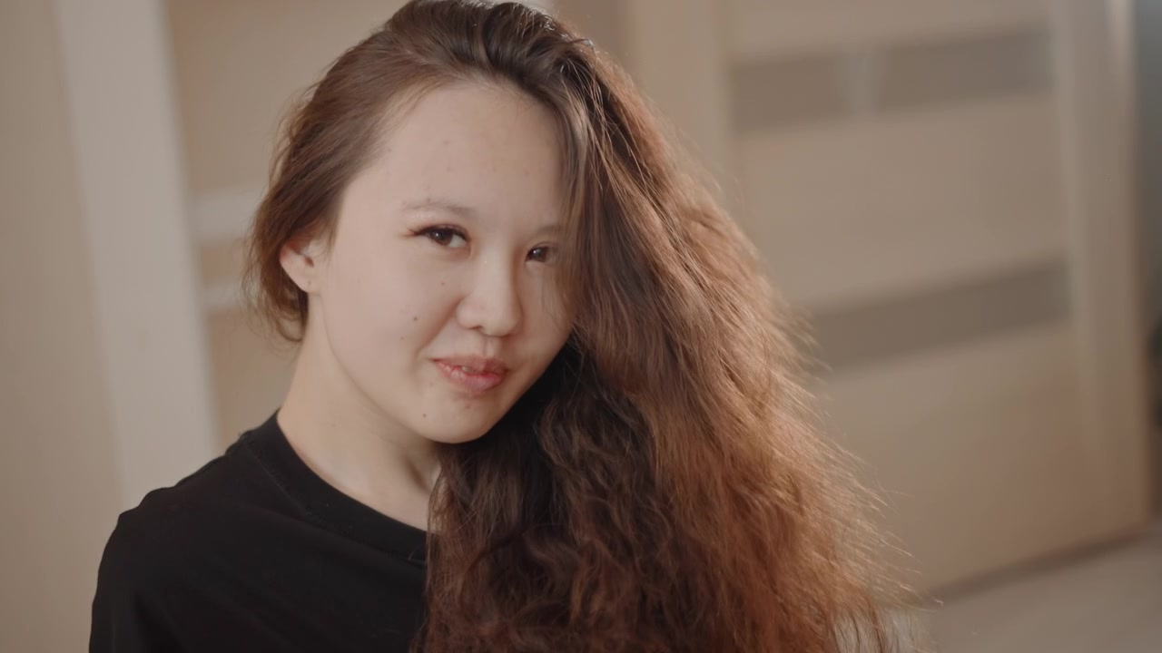 長い赤褐色の巻き毛の若い女性が、髪に手を通しながらカメラの方を向いている。髪にボリュームが出て、流れ落ちる髪の房、柔らかな笑顔、自信に満ちたアイコンタクトが、ニュートラルな室内光の中で際立つ。