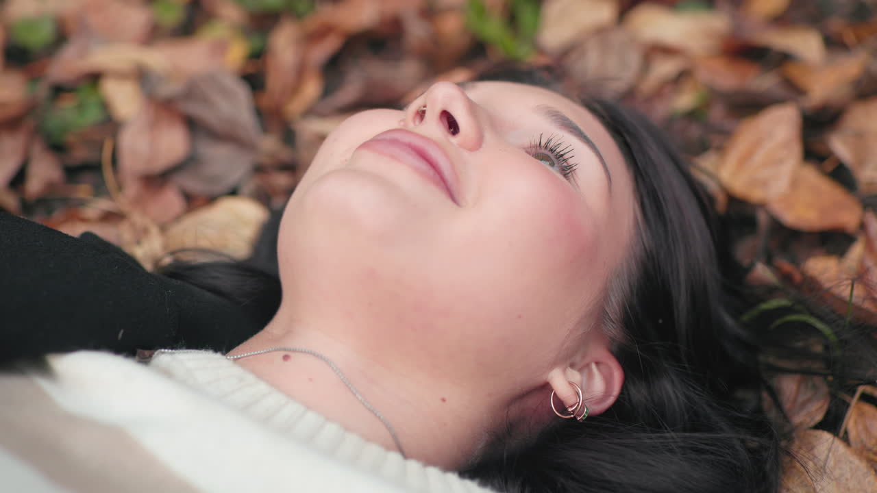 Close zoom on autumn lover lying on leaf carpet, eyes closed, laughing out loud as single dry leaf falls beside face, cozy sweater and black coat, warm forest background, carefree seasonal joy