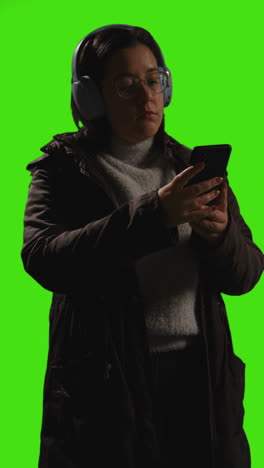 Vertical-Video-Of-Woman-Wearing-Wireless-Headphones-Streaming-Music-From-Mobile-Phone-Against-Green-Screen-With-Low-Key-Lighting