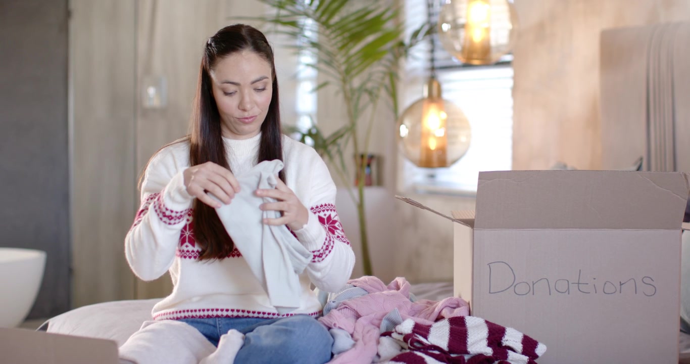 女性がベッドの上でグレーのセーターを確認し、畳んでクリスマス用の寄付箱に入れている。