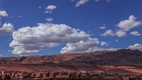 time lapse shot of flying white clouds on blue sky over red mountains and small african city in desert of morocco