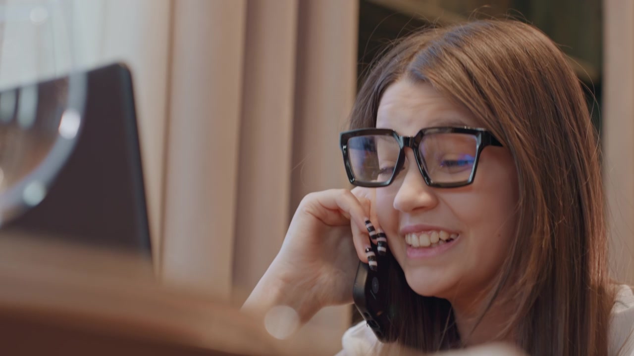 Young woman on phone looking worried, glasses and laptop at small desk, heavy eyelids and occasional yawn, late study session interrupted by billing or support issue, quiet indoor atmosphere