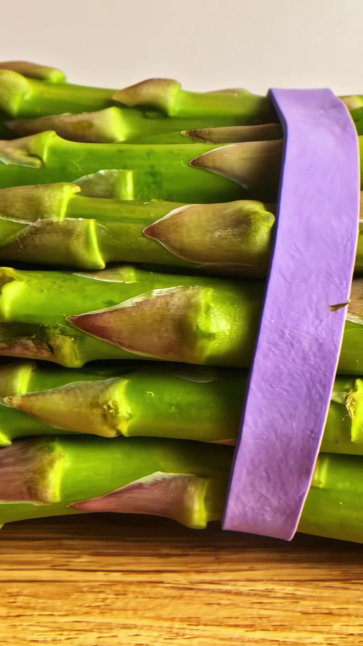 Un mazzetto di asparagi verdi freschi legato con un elastico viola su un tagliere di legno, primo piano con carrello a destra