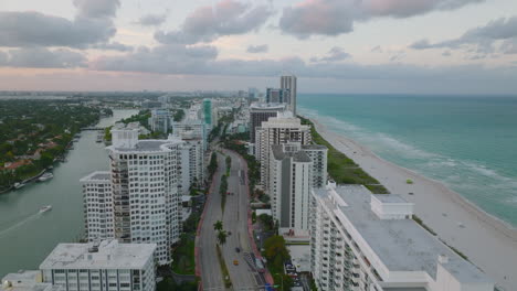 Fly-above-multistorey-buildings-at-sea-coast-at-dusk.-Cars-driving-on-multilane-road-between-apartment-houses.-Miami,-USA