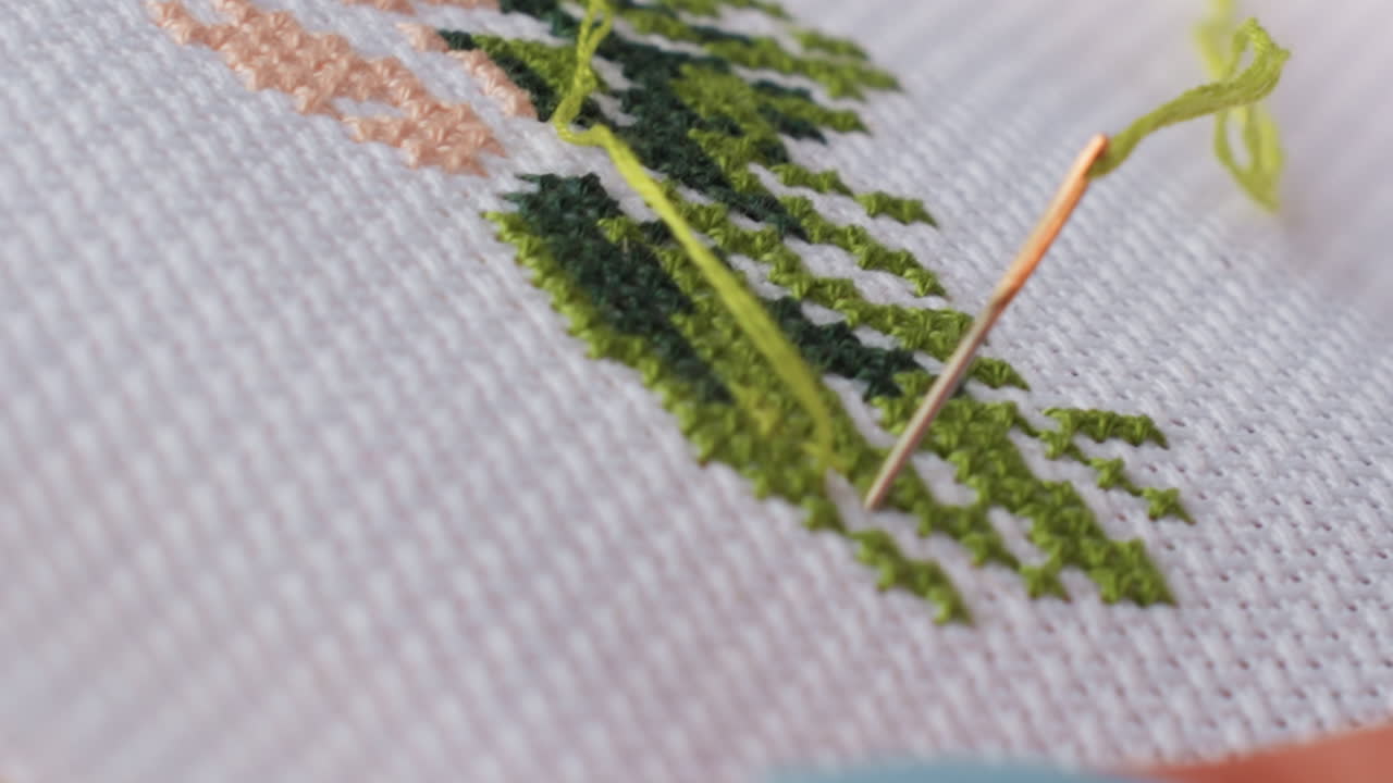Extreme close up shows crafter passing needle with green thread through cross stitch fabric, shaping leafy pattern on white canvas, crisp weave and texture visible