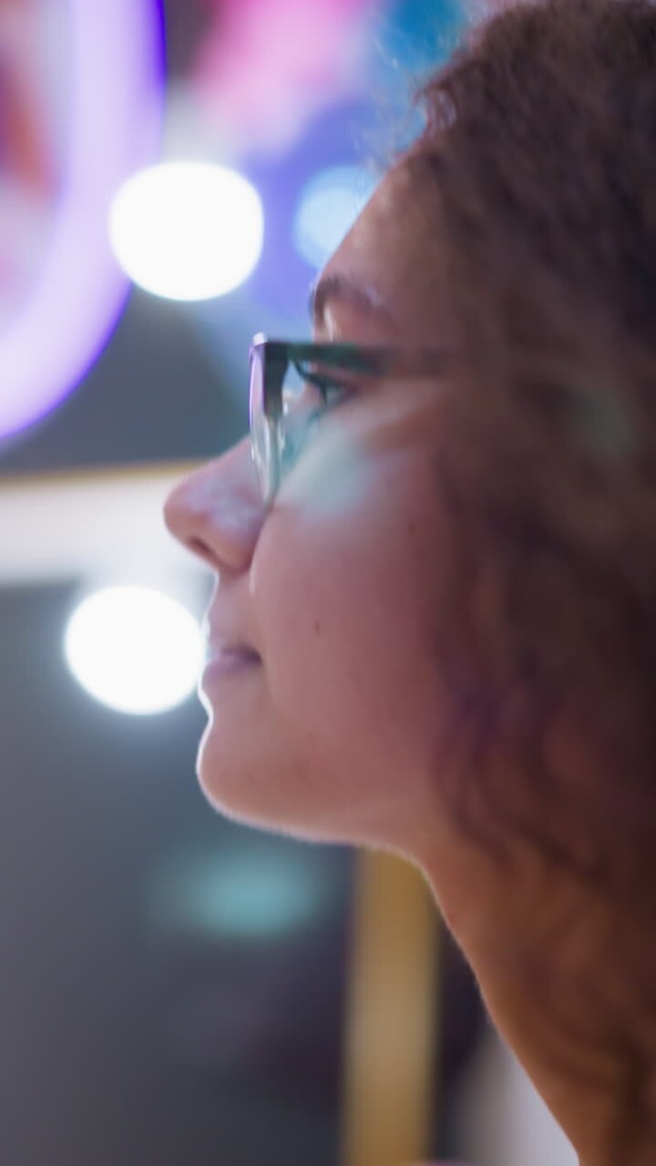 Lady with curly hair and glasses walking through well-lit mall, displaying a calm and focused expression, blurred background reveals bustling retail environment with stores and people