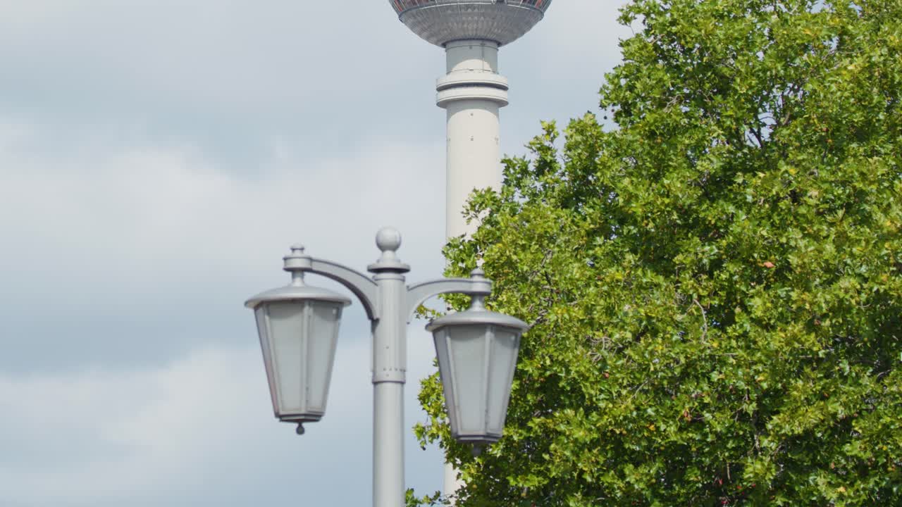 Camera tilts upward revealing TV tower, streetlights, green trees, and cloudy blue sky in Berlin