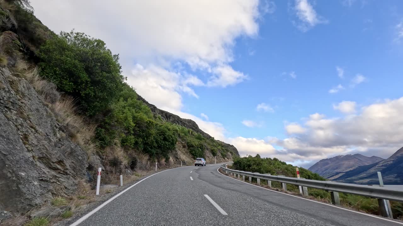 Forward-facing car dashcam captures winding mountain road, lakeside scenery, and passing vehicles in daylight
