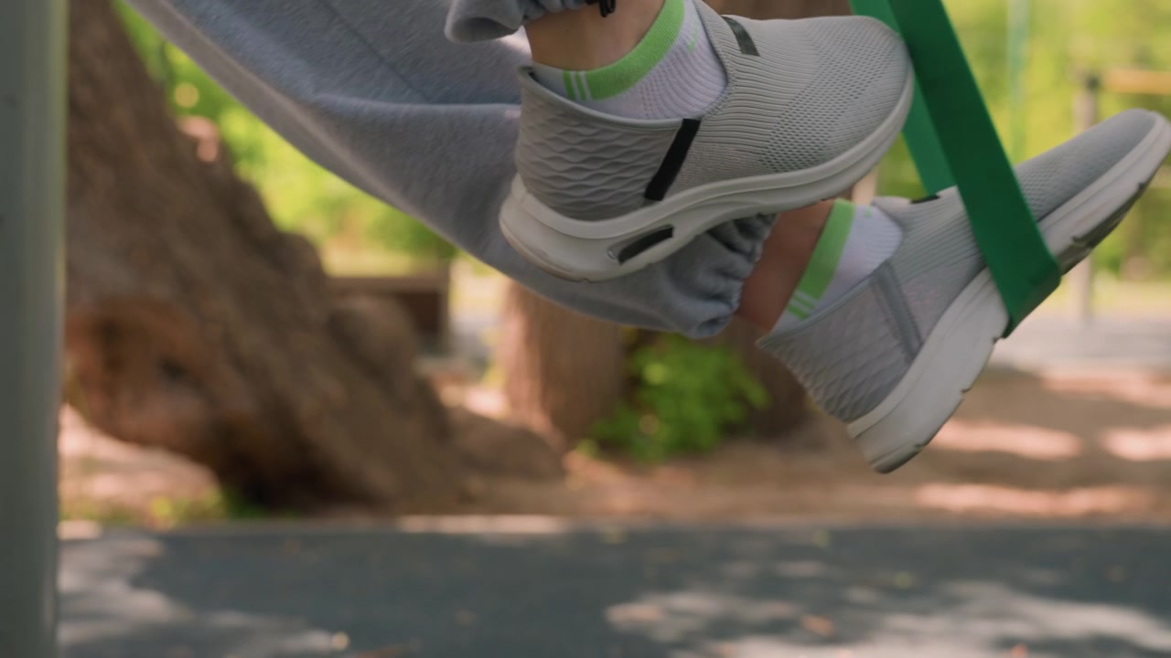 Primo piano delle gambe e dei piedi di una persona che esegue esercizi con elastici di resistenza all'aperto. Il soggetto indossa pantaloni della tuta grigi, calzini bianchi con accenti verdi e scarpe sportive grigio chiaro mentre usa l'elastico