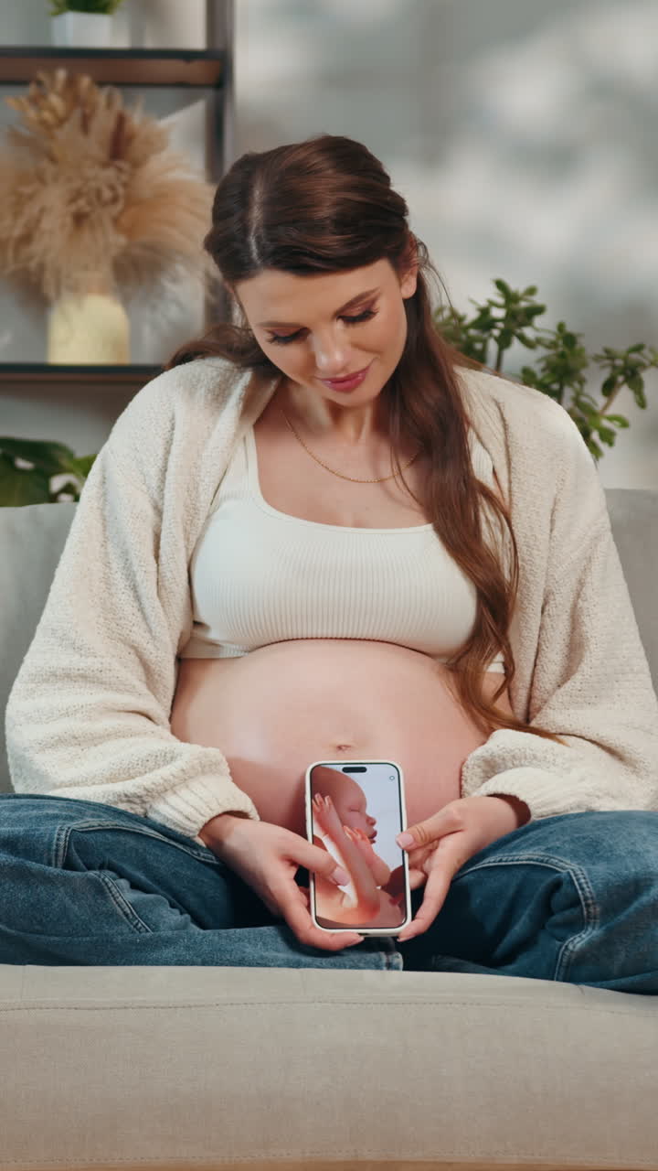 Pregnant woman sits on couch holding phone displaying ultrasound image of babys feet anticipating