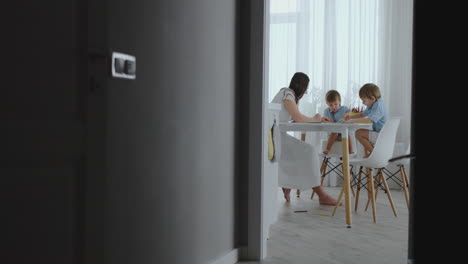 mom helps sons learn to draw doing homework preschool preparation at home sitting in the white kitchen. two children brothers draw a portrait of mother together