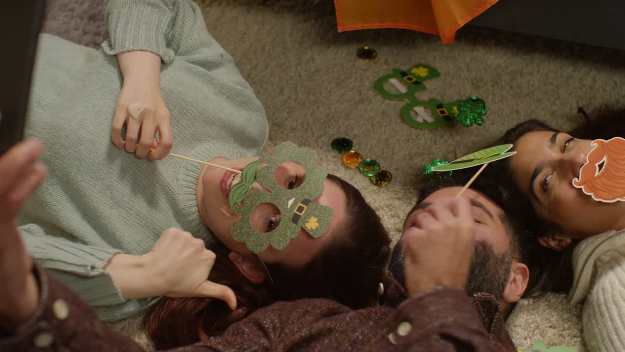 группа друзей одевается дома, лежит на полу, празднует день святого патрика, позирует для селфи на телефоне.