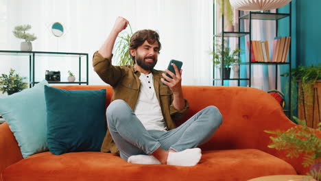 Middle-eastern-young-man-at-home-use-smartphone-celebrating-success-victory-winning-play-online-game