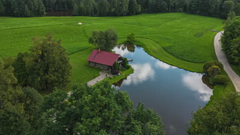 a smooth aerial shot of a house by a pond at a scenic green landscape near a grove