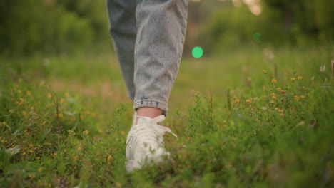 close-up of legs in white sneakers and rolled-up jeans, walking through a grassy field with patches of brown foliage, blurred red and green lights appear in the distant background