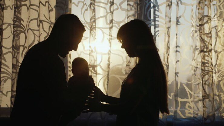 Silhouette Of Mom And Dad Sitting By The Window At Sunset They Play With Their Baby Happy Family At