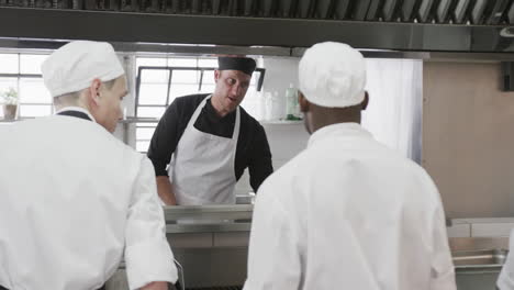 Focused-diverse-male-chef-instructing-trainee-male-chefs-in-kitchen,-slow-motion