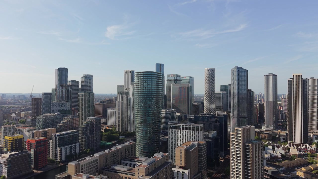 Numerosos edificios modernos y altos dominan el distrito financiero de Canary Wharf en Londres. Los rascacielos se elevan hacia un cielo azul claro. La brillante luz del sol ilumina el denso paisaje urbano durante el día
