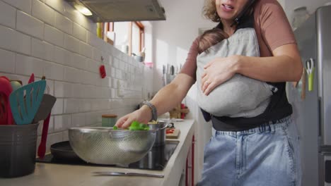 Video-of-caucasian-mother-with-newborn-baby-in-baby-carrier-using-smartphone-in-kitchen