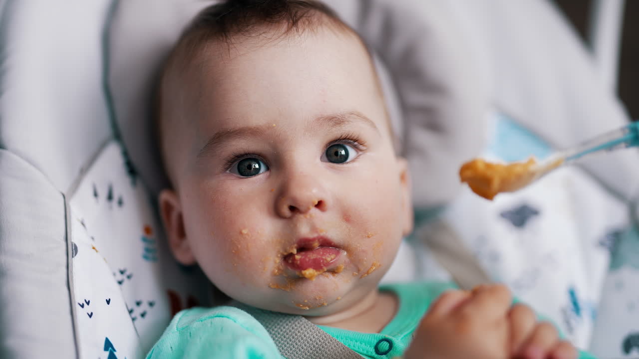 Feeding beautiful child from spoon. Baby looks ahead and then smiles and waves hands cheerfully. Close up.