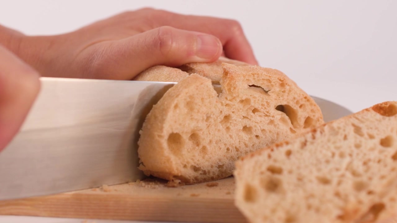 Hands use a sharp chef knife to slice artisan sourdough bread on a wooden board.